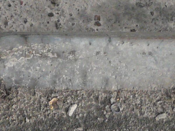 Road edge texture, with uneven grey asphalt and a cement area with a swirling discoloration, divided by a strip of similarly colored stone.